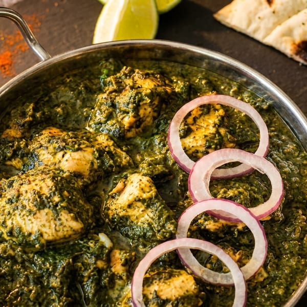 Palak chicken in a metal bowl with red onion rings, lime, naan, and a spoon.