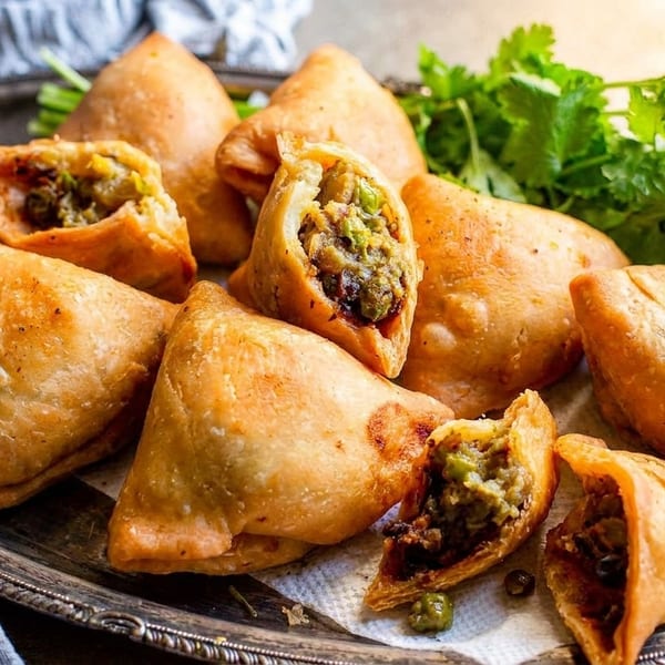 Golden-brown samosas, some broken open to show the savory filling, served on a silver platter with green chutney.
